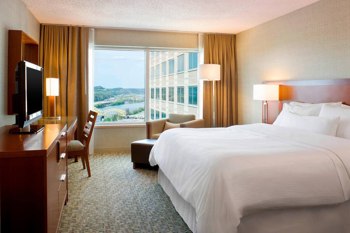 A hotel room with a large bed, a desk with a TV, a chair, and a view of a bridge and greenery.