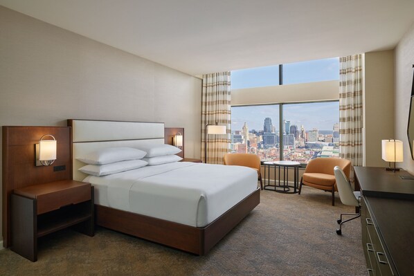 Una habitación de hotel con una cama grande, dos mesitas de noche con lámparas, una pequeña mesa con una silla y vistas a un paisaje urbano a través de amplias ventanas.