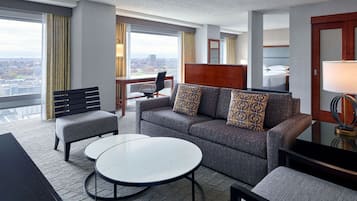 A modern hotel room with a gray sofa, two round tables, a desk, and a large window offering a city view.
