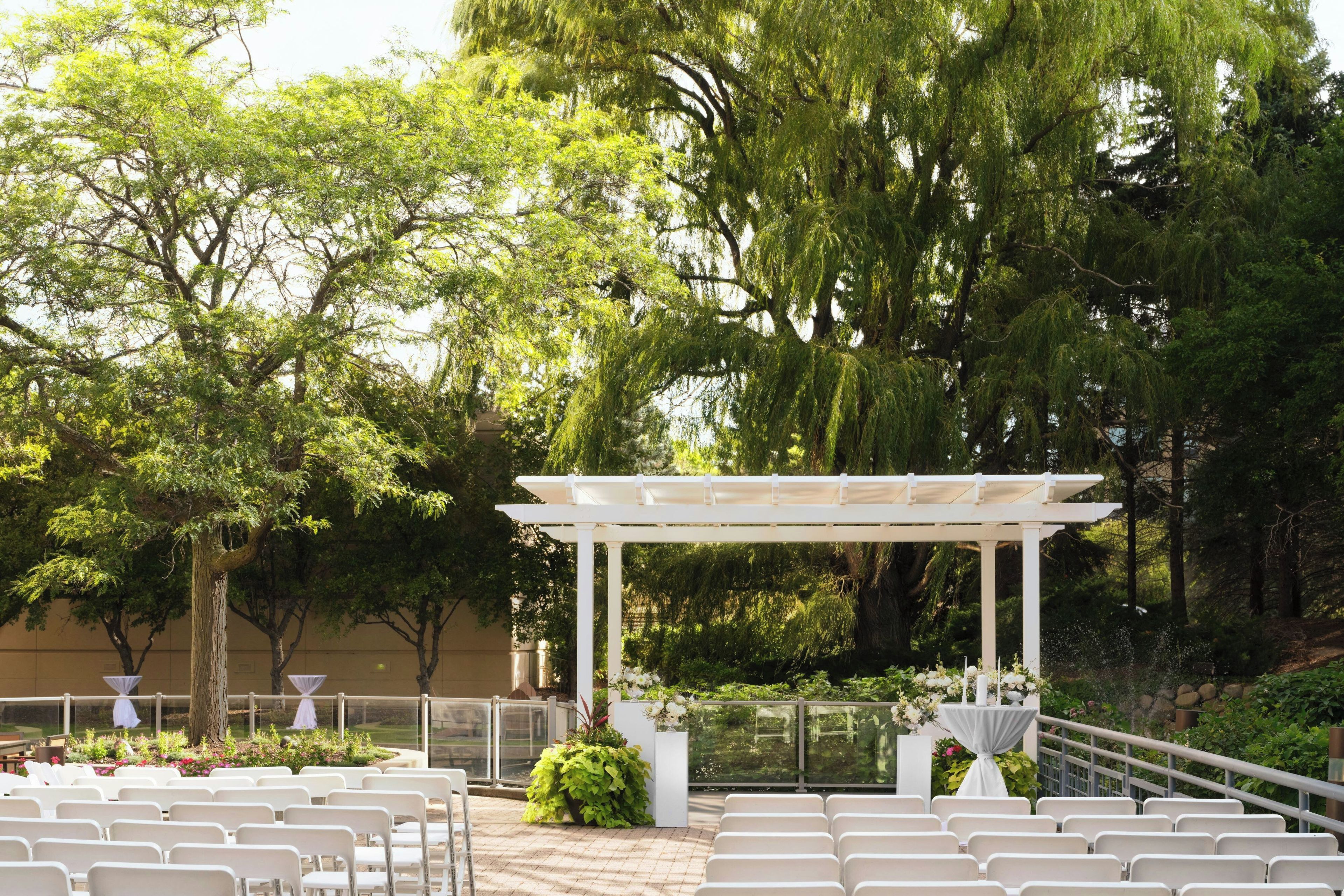 Un espacio para bodas al aire libre con una pérgola blanca, filas de sillas blancas y abundante follaje de fondo.