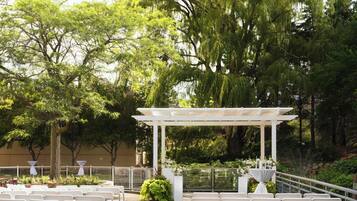Un espacio para bodas al aire libre con una pérgola blanca, filas de sillas blancas y abundante follaje de fondo.