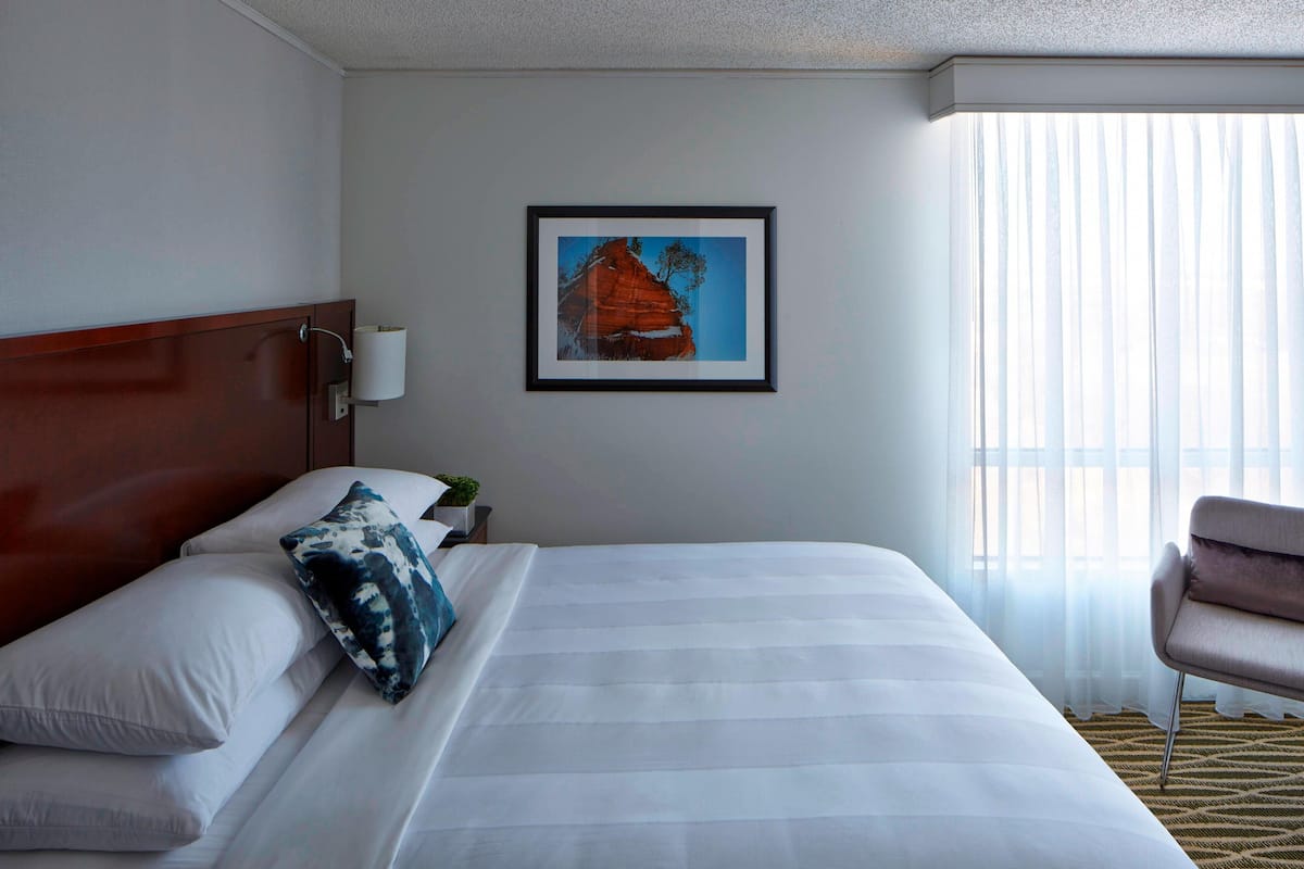 Une chambre d’hôtel avec un grand lit, une tête de lit, un tableau encadré au mur, une chaise et une fenêtre avec des rideaux légers.