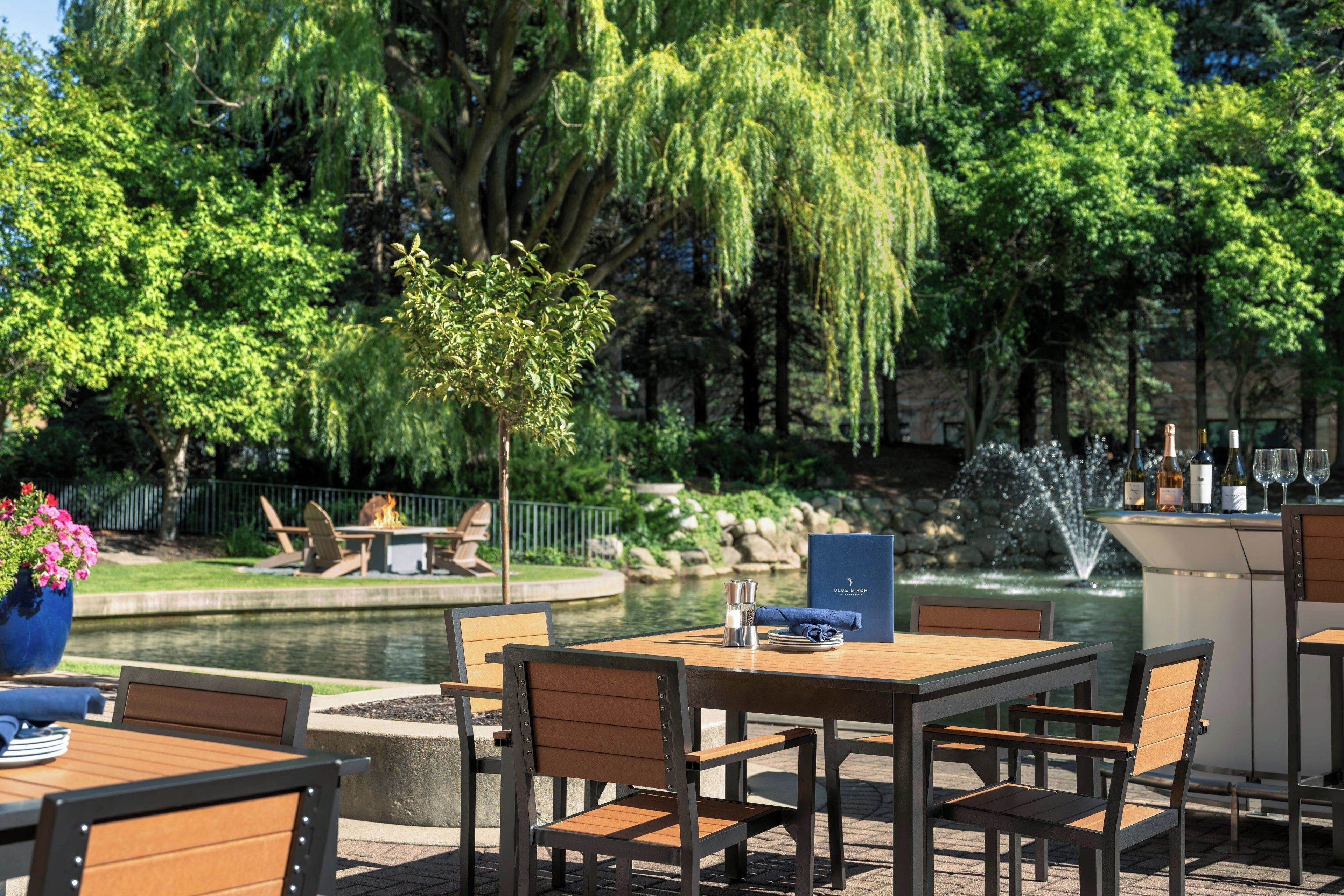 An outdoor seating area with tables and chairs, a small tree, and a fountain.