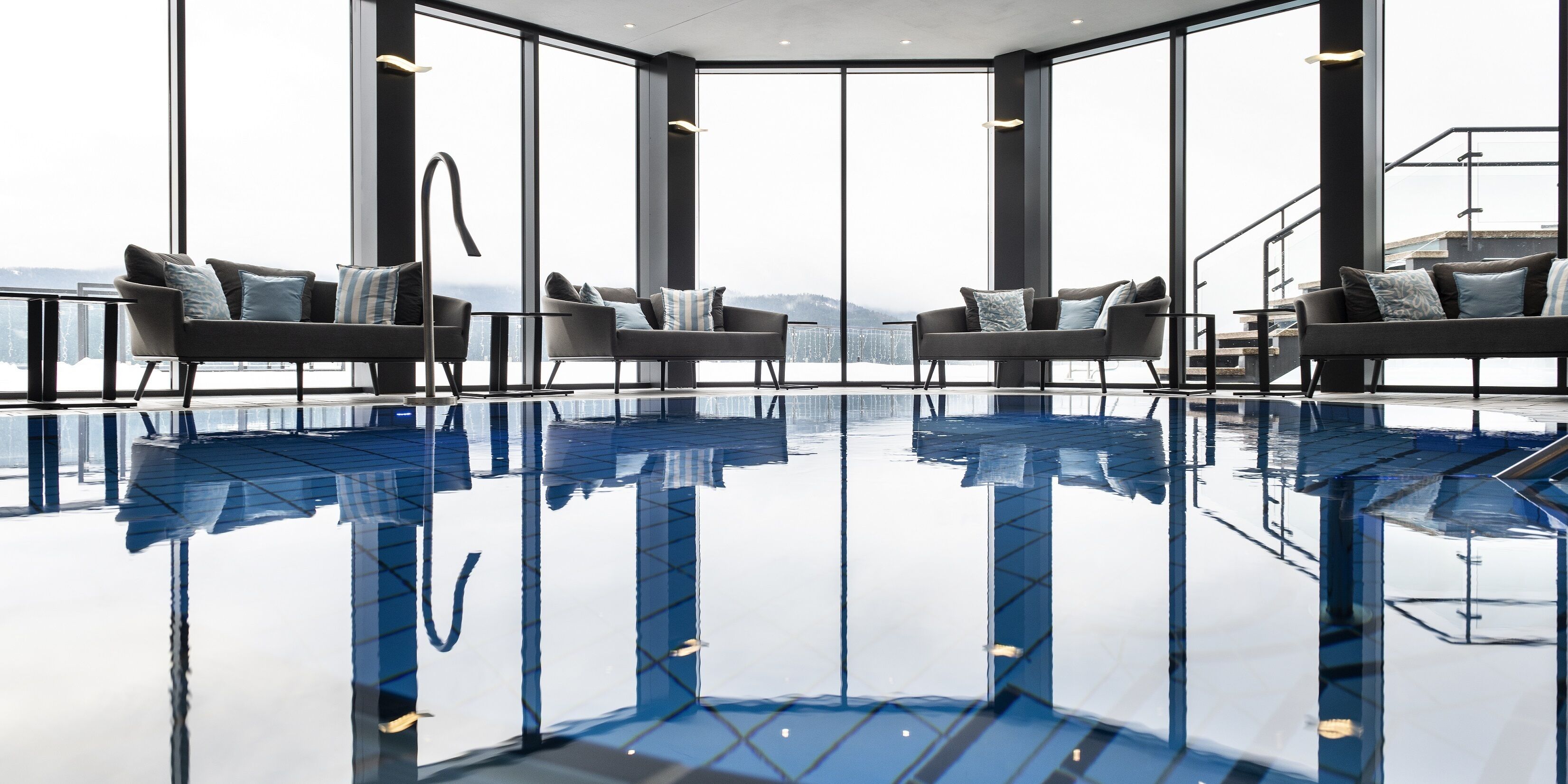 Ein modernes Interieur mit einem großen, spiegelnden Pool, zwei Sofas mit Kissen und großen Fenstern mit Blick auf die Stadtlandschaft.