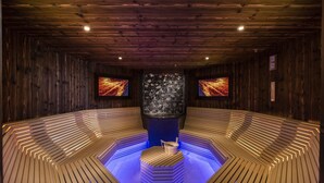 Uma sauna com bancos de madeira dispostos em semicírculo, uma unidade de aquecimento central e duas telas fixadas na parede exibindo imagens vibrantes.