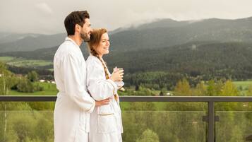 A couple in white robes standing on a balcony with a view of a green valley and mountains.