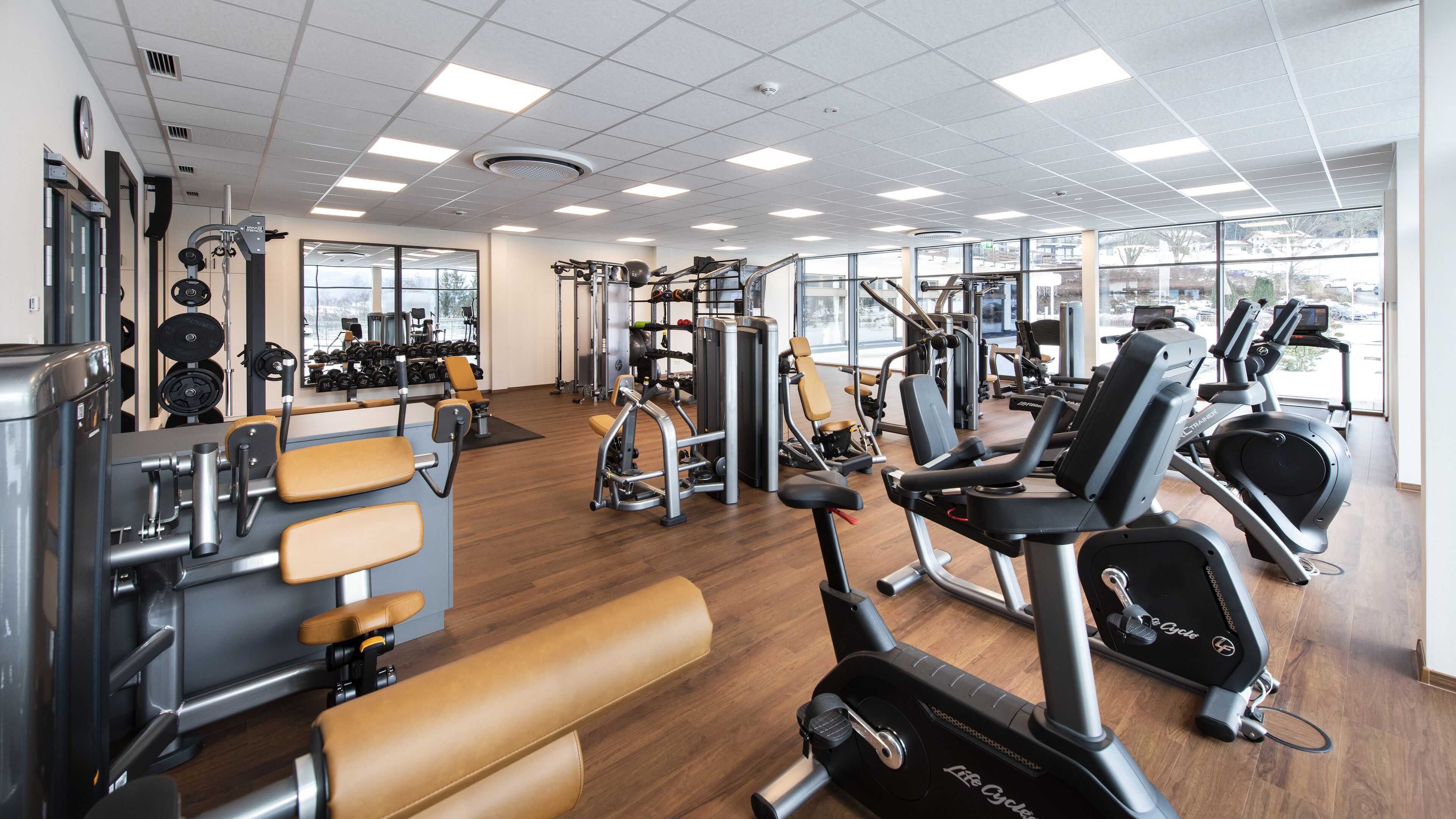A gym with various exercise equipment, large windows, and a wooden floor.