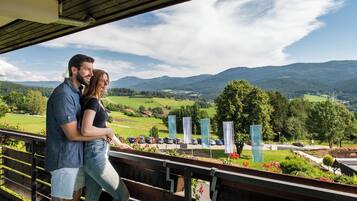 Ein Paar, das sich auf einem Balkon bei einem malerischen Ausblick auf eine Berglandschaft, einen Parkplatz und einen Blick auf ein Dorf umarmt.