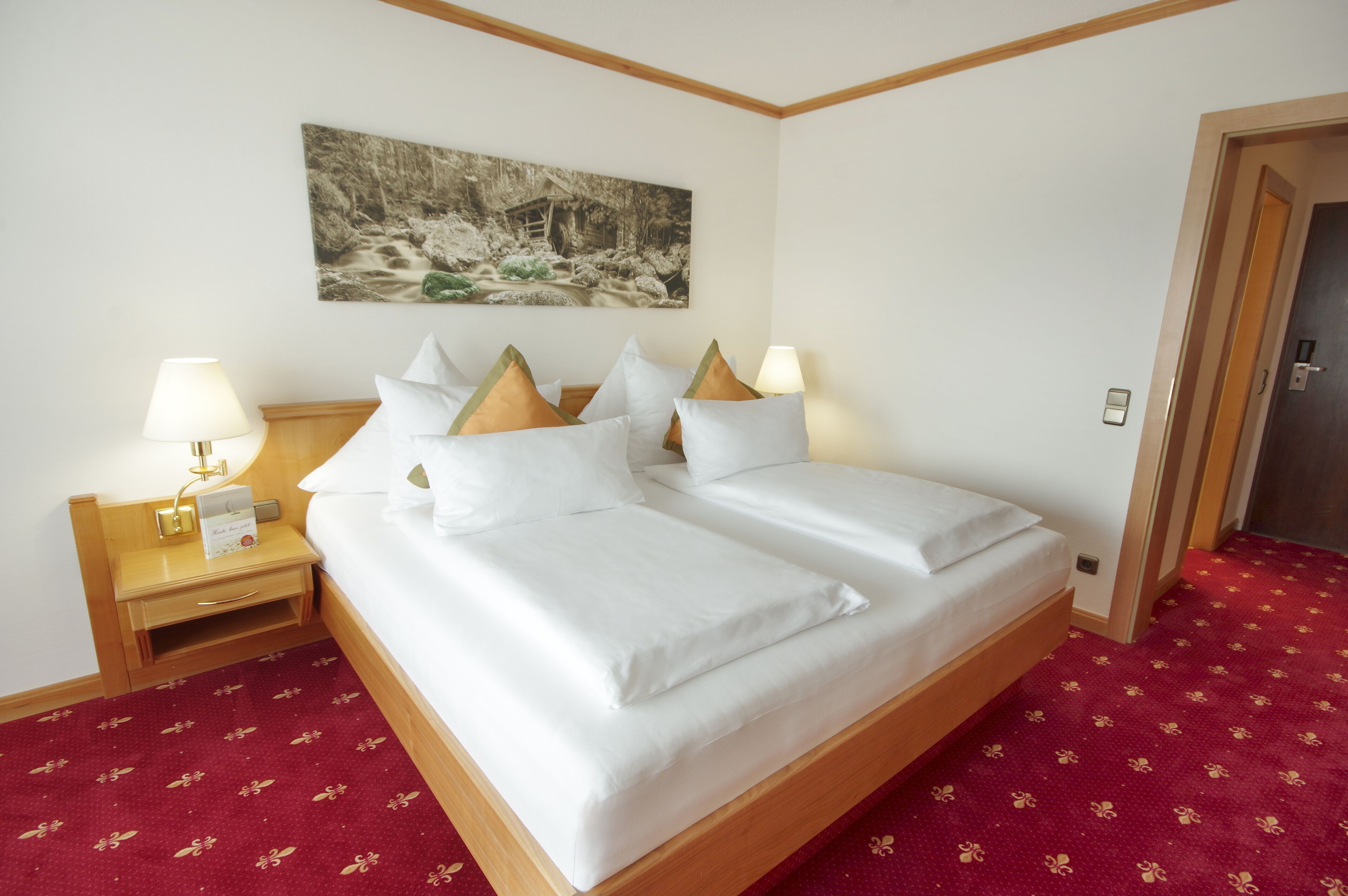 A neatly made bed with white linens and a wooden headboard, two bedside tables with lamps, a red patterned carpet, and a painting of a forest scene on the wall.