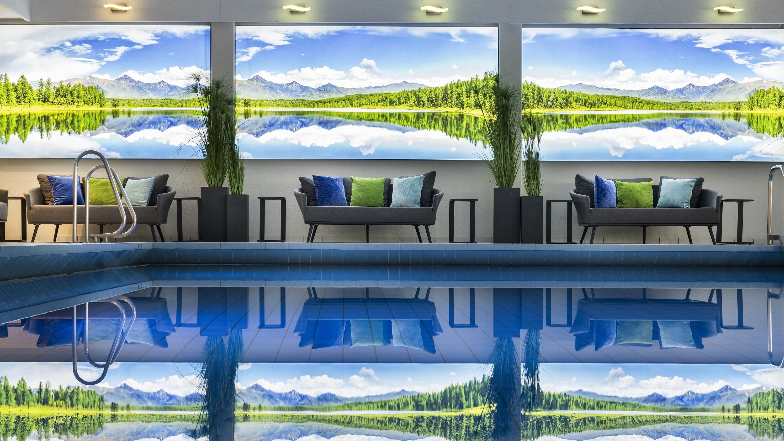Ein Poolbereich mit Blick auf Berge und einen See durch große Fenster. Zwei Sofas mit Kissen und Topfpflanzen sind vorhanden.