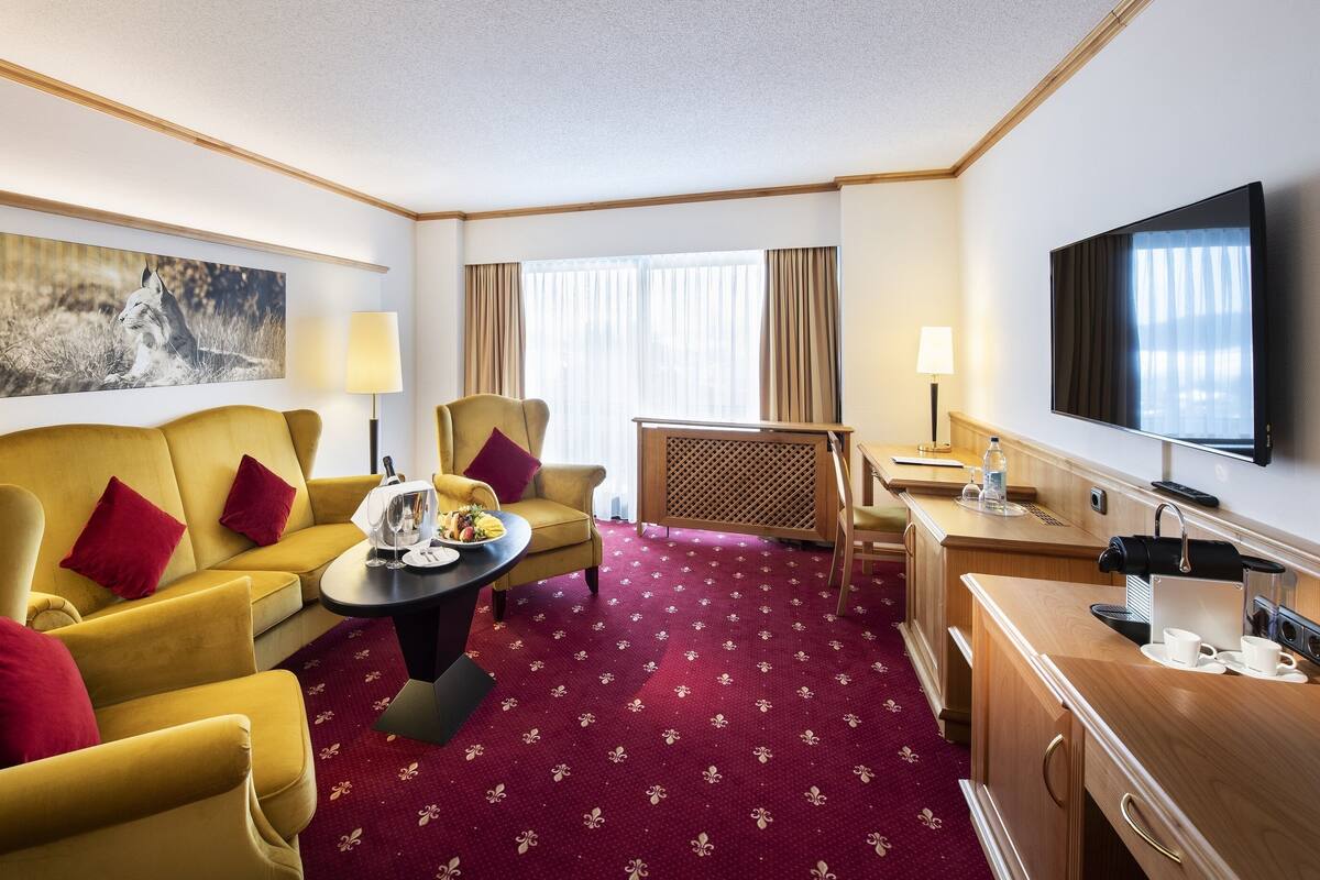 A hotel room with a yellow sofa, red cushions, a round coffee table, a TV, a desk with a lamp, and a large window with curtains.