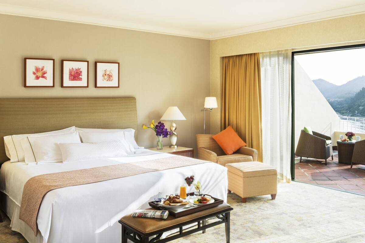 A hotel room with a large bed, a sitting area with a chair and a table, and a balcony with chairs.