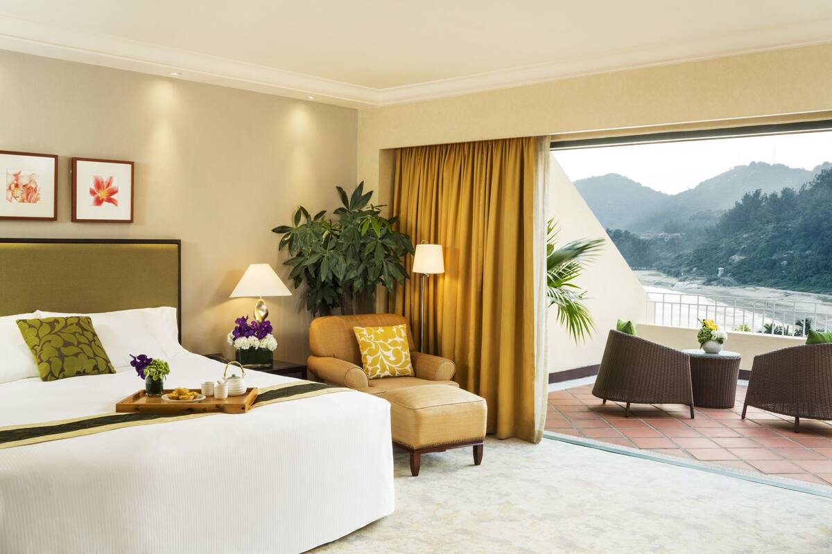 A hotel room with a bed, a chair, a table, a plant, and a view of a mountainous landscape.