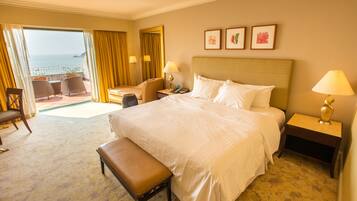 A hotel room with a large bed, a bench, a desk, and a chair. There is a balcony with a view of the sea.