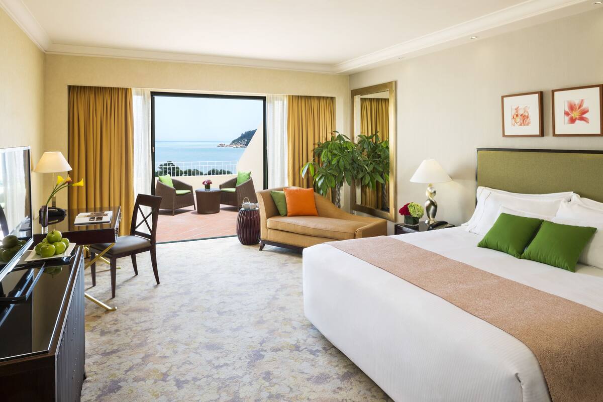A hotel room with a large bed, a desk with a lamp, a chair, a plant, and a view of the ocean.