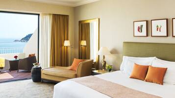 Habitación de hotel con cama, sofá, sillón, mesita de noche, lámpara, ventana con cortinas y cuadros en la pared.
