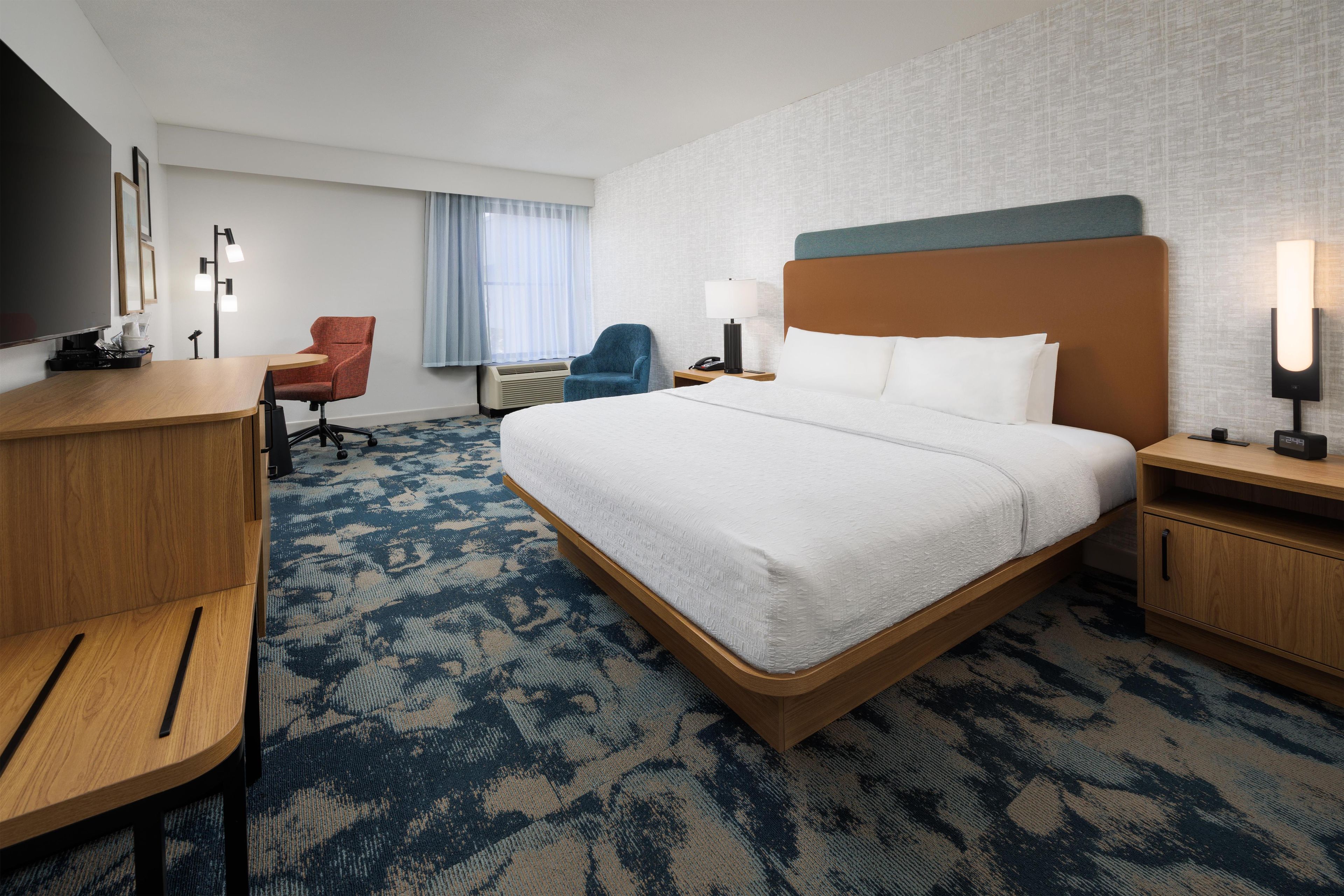 Quarto de hotel com uma cama grande, uma escrivaninha, uma cadeira, uma televisão e um abajur.