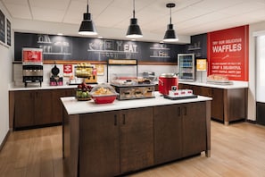 A modern kitchen area with a central island, a juice dispenser, a waffle display, and a breakfast sign.