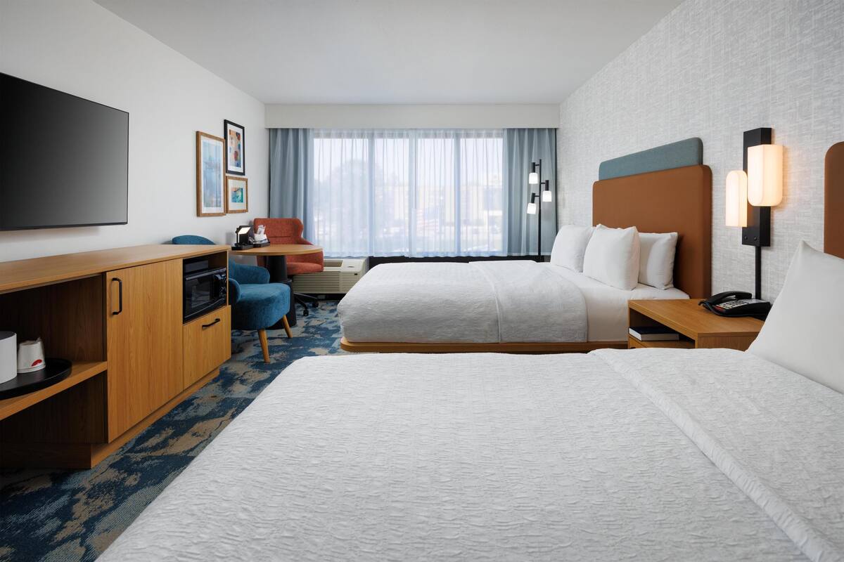 A hotel room with two beds, a desk, a TV, and a window with curtains.