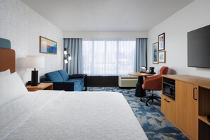 Una habitación de hotel con cama, escritorio, silla, sofá, televisor y un ventanal con cortinas.