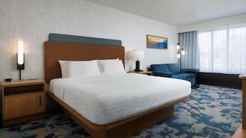 A hotel room with a large bed, a wooden nightstand, a desk, a blue armchair, and a window with curtains.