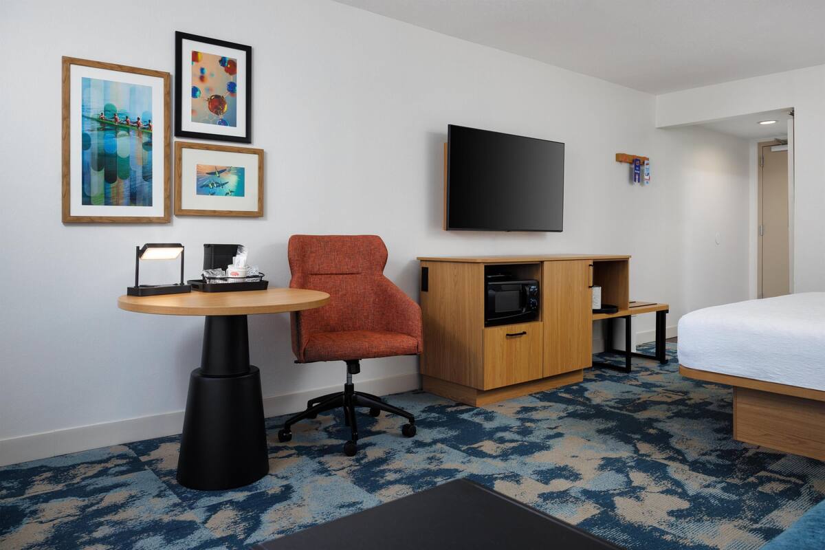 A hotel room with a bed, a TV, a desk with a chair, a microwave, and framed pictures on the wall.