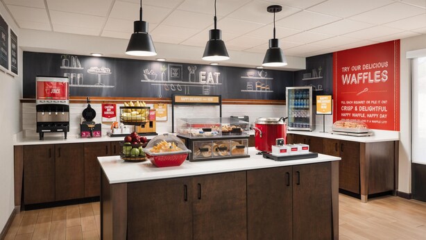 A modern kitchen area with a central island, a juice dispenser, a waffle display, and a breakfast sign. - Hampton Inn Oak Ridge Knoxville (Oak Ridge)