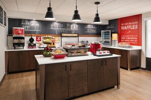 A modern kitchen area with a central island, a juice dispenser, a waffle display, and a breakfast sign.