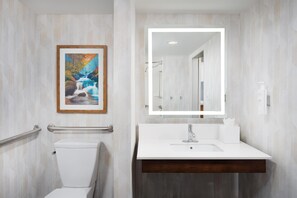 A bathroom with a white sink, a framed painting, a mirror with lights, and a handrail.