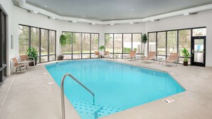 A large indoor swimming pool with clear blue water, surrounded by large windows offering views of the outdoors. There are several lounge chairs and a small table with chairs around the pool. - Hampton Inn Oak Ridge Knoxville (Oak Ridge)