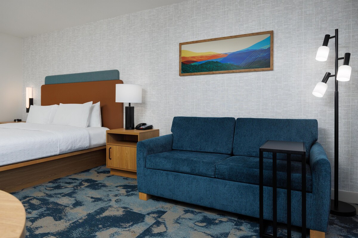 Una habitación de hotel con cama, sofá azul, mesita de noche con lámpara y un cuadro en la pared.