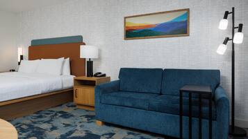 A hotel room with a bed, a blue sofa, a nightstand with a lamp, and a painting on the wall.