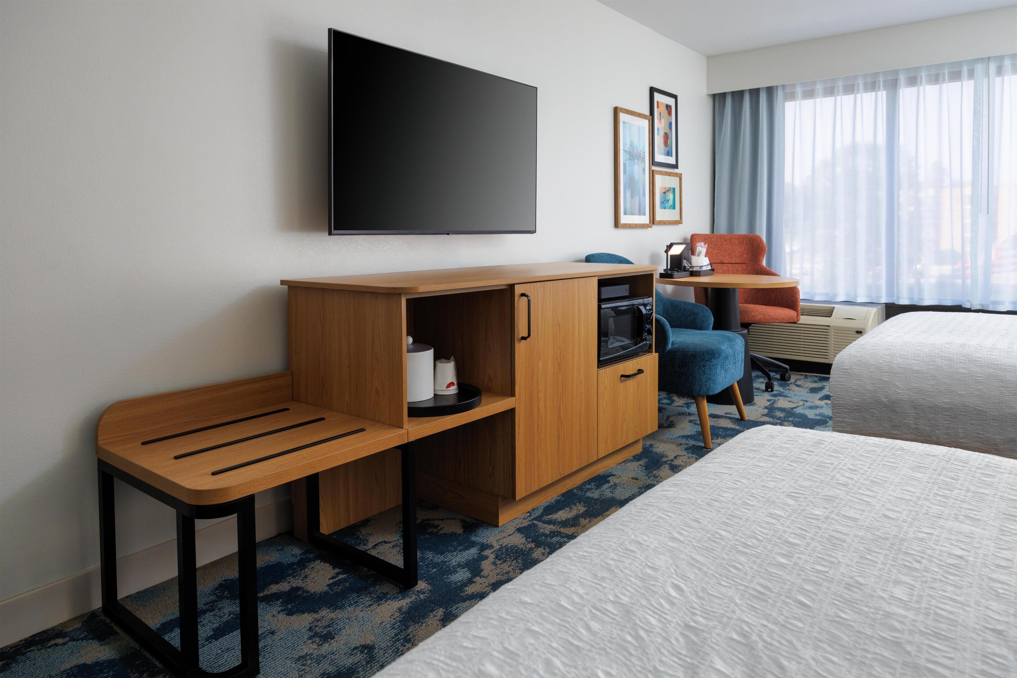 Une chambre d’hôtel comprenant un lit, un bureau avec un téléviseur à écran plat, un coin salon avec un fauteuil et une fenêtre avec des rideaux.