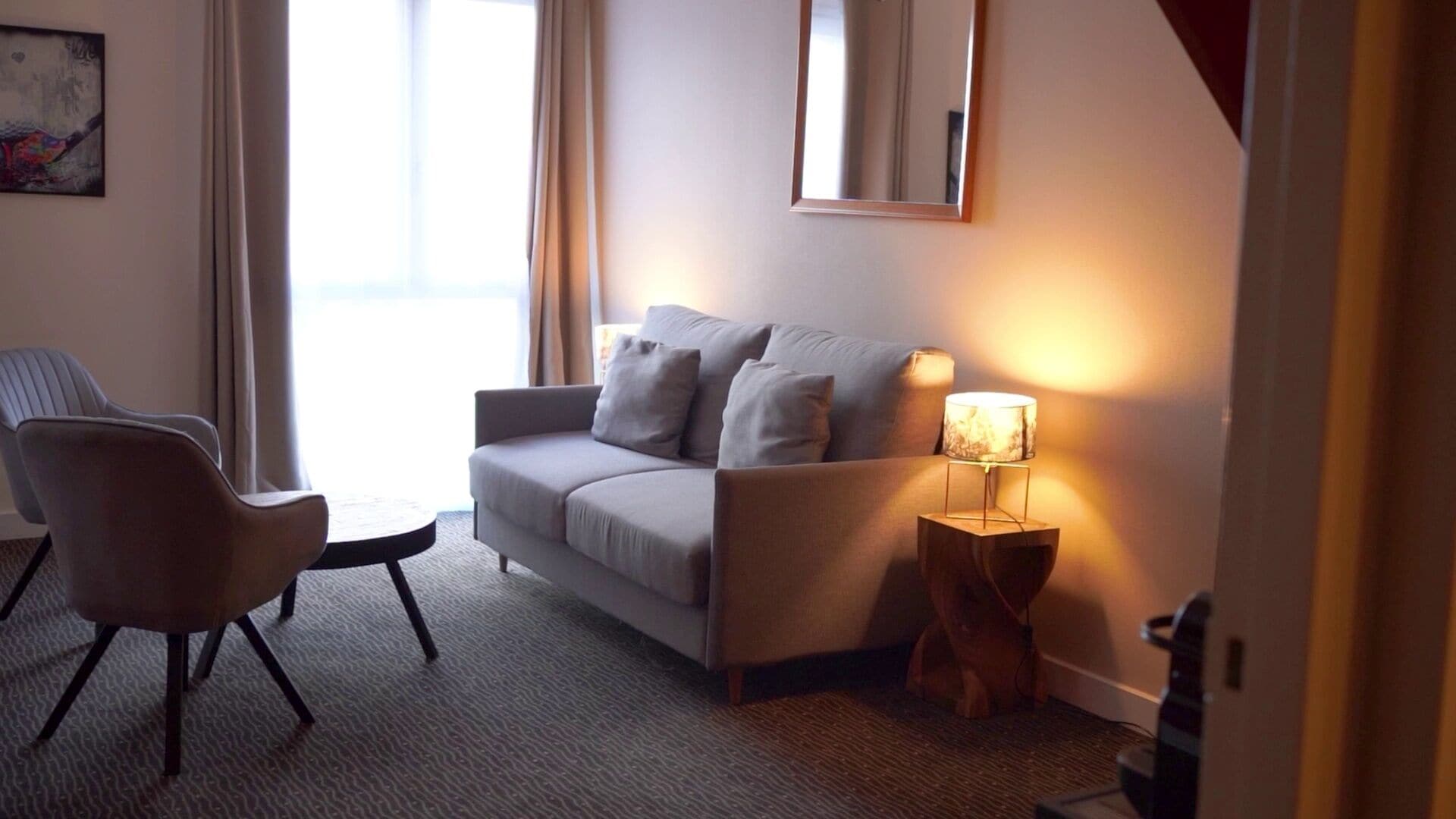Ein modernes Hotelzimmer mit grauem Sofa, einem einzelnen Sessel, einem kleinen runden Tisch und einer Lampe auf einem Holzfuß.