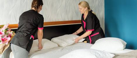 Two individuals arranging pillows on a bed in a hotel room with a textured wall and a wooden headboard.