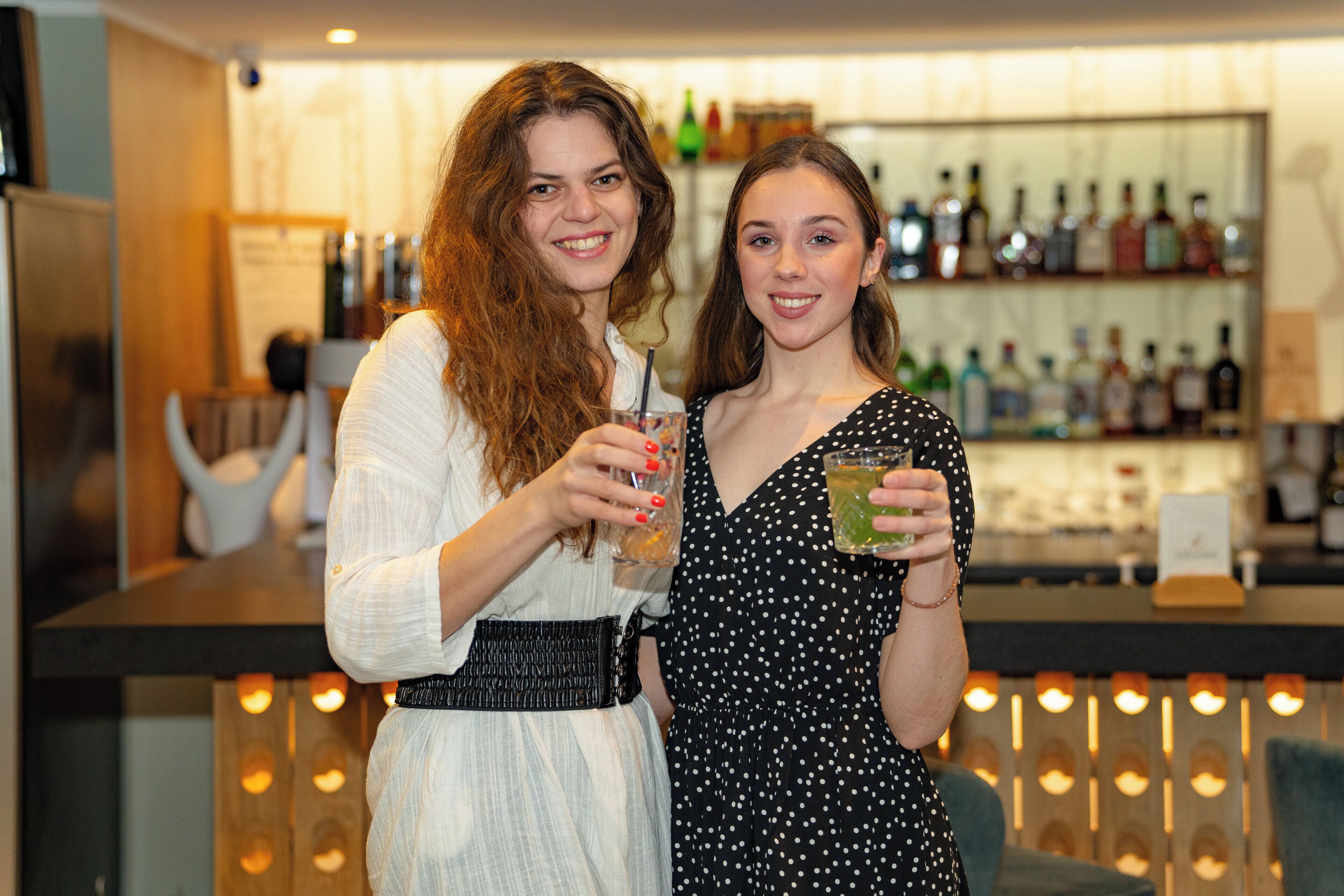 Deux personnes sont debout au bar, et on peut voir une variété de bouteilles en arrière-plan.