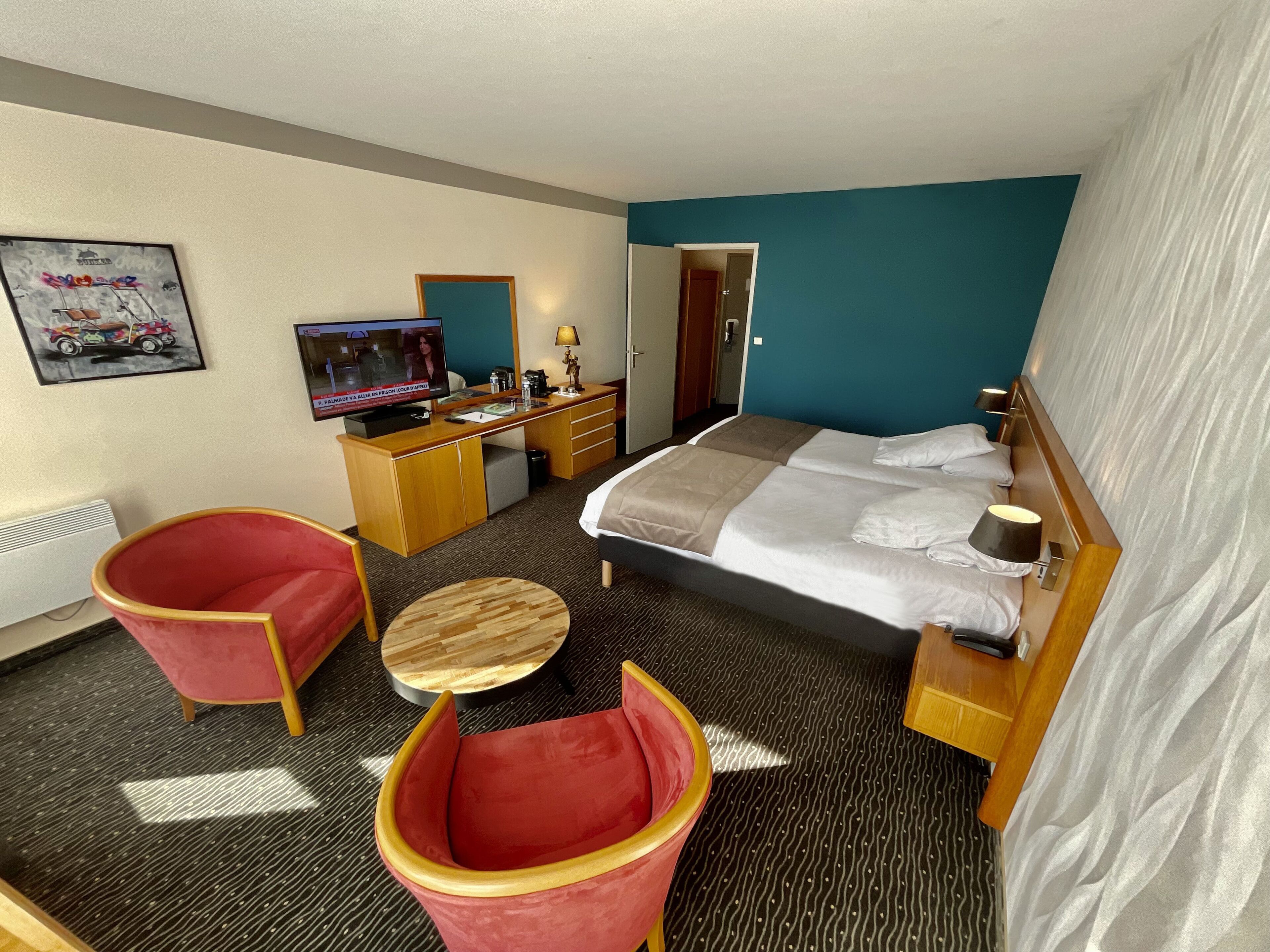 Une chambre d’hôtel avec un lit, un bureau, une télévision, une chaise rouge et une petite table ronde.