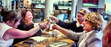 A group of people toasting with wine glasses at a table in a restaurant with a bar in the background.