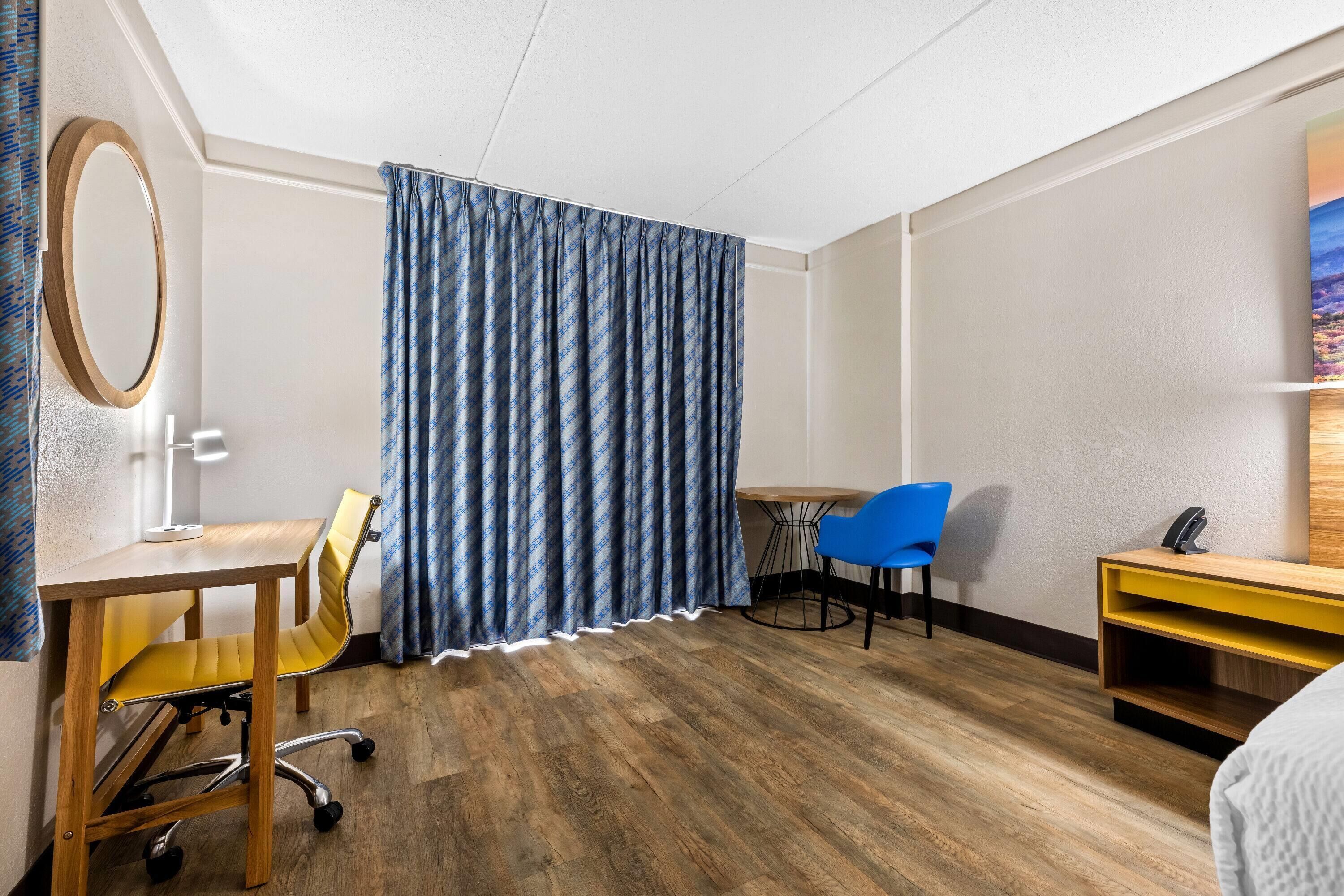 Habitación de hotel con un escritorio de madera, una silla, una mesita pequeña, una cortina azul y un cuadro en la pared.