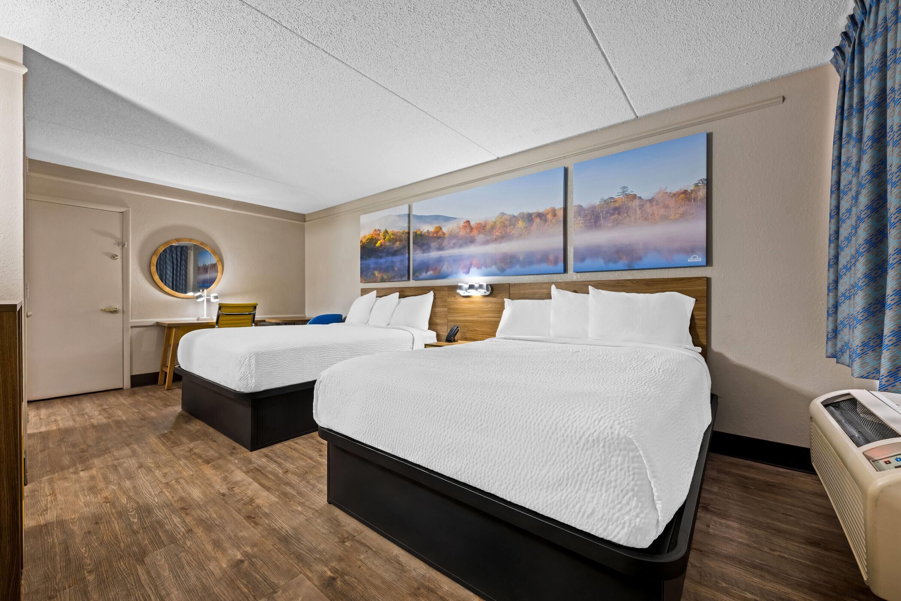 A hotel room with two beds, a round mirror, a nightstand, a wall-mounted TV, and a large window with a view of autumn foliage.