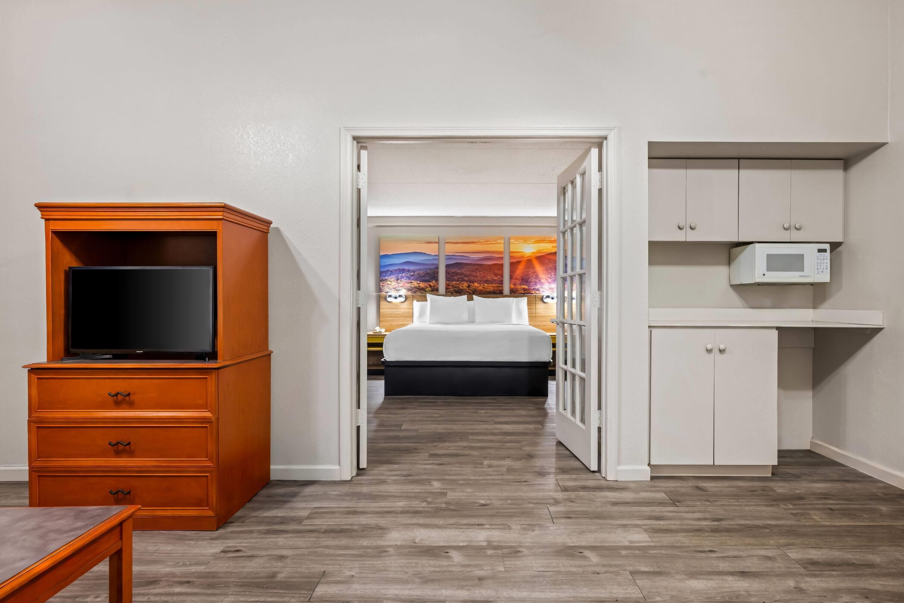 Une chambre d’hôtel avec un meuble en bois pour la télévision, un lit avec une literie blanche, un four à micro-ondes et une vue sur le coucher du soleil par la fenêtre.