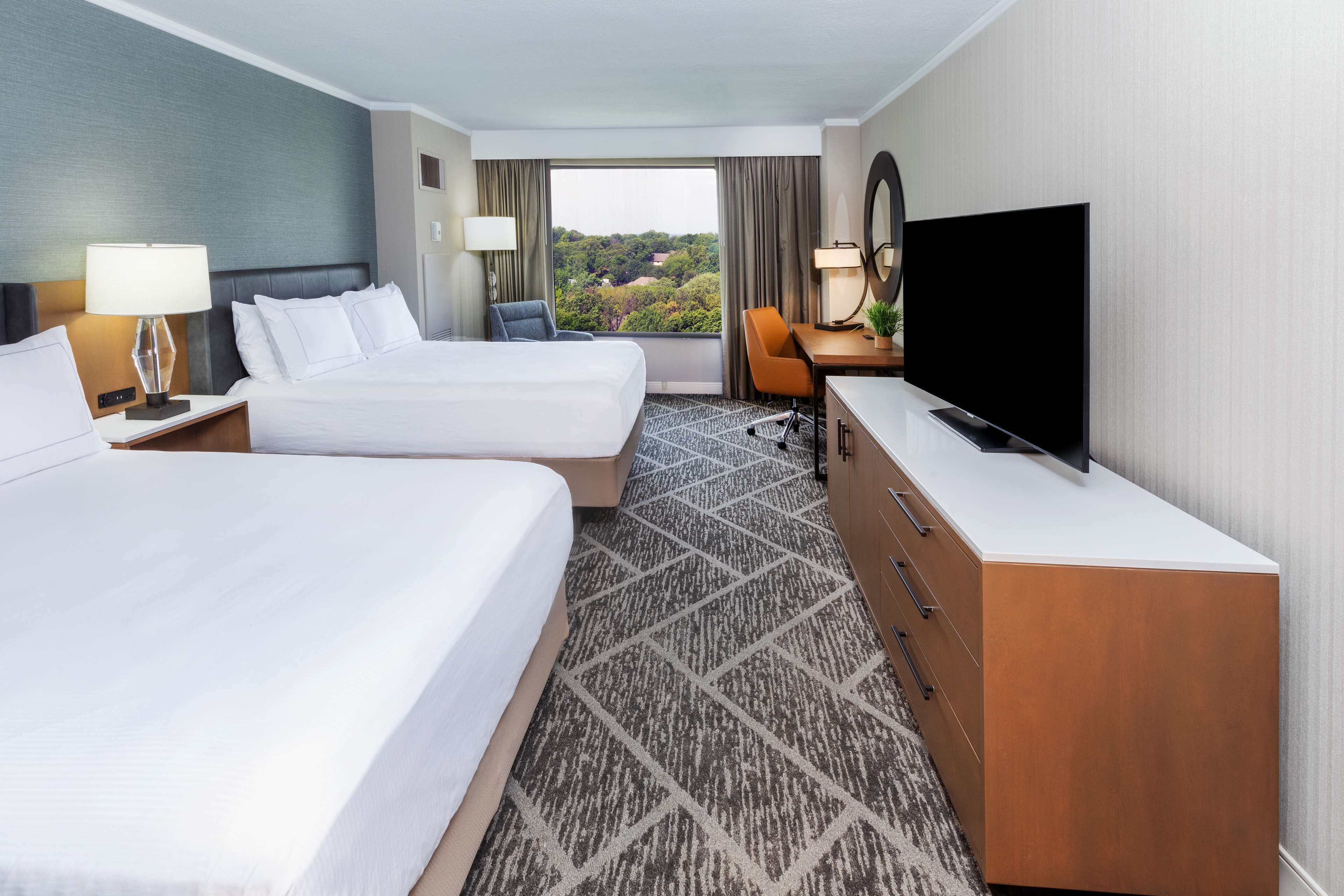 A hotel room with two beds, a TV, a desk, and a view of the outdoors.