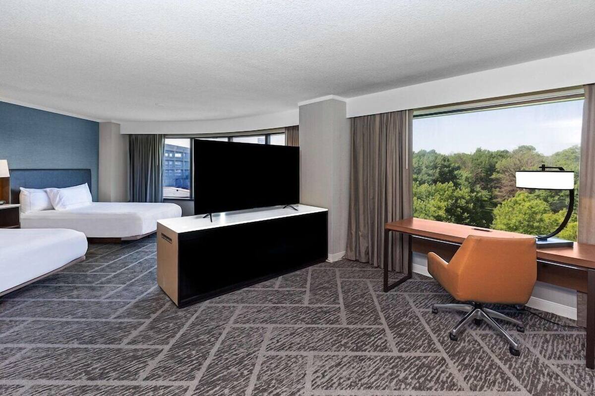 A hotel room with a bed, a desk, a chair, a TV, and a window with a view of trees.
