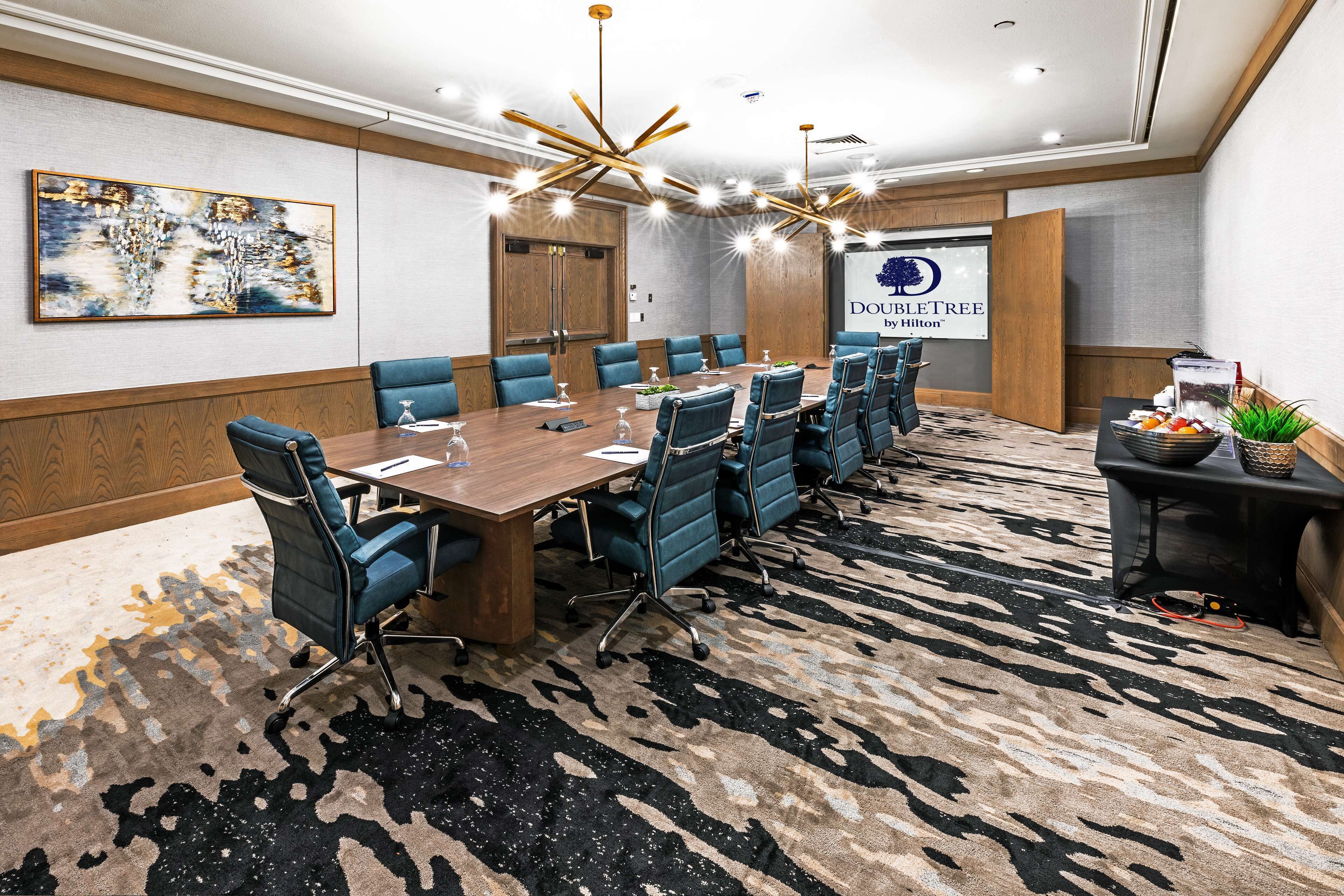 Un salón de conferencias con una larga mesa, sillas azules y una pantalla grande que muestra el logotipo de DoubleTree by Hilton.