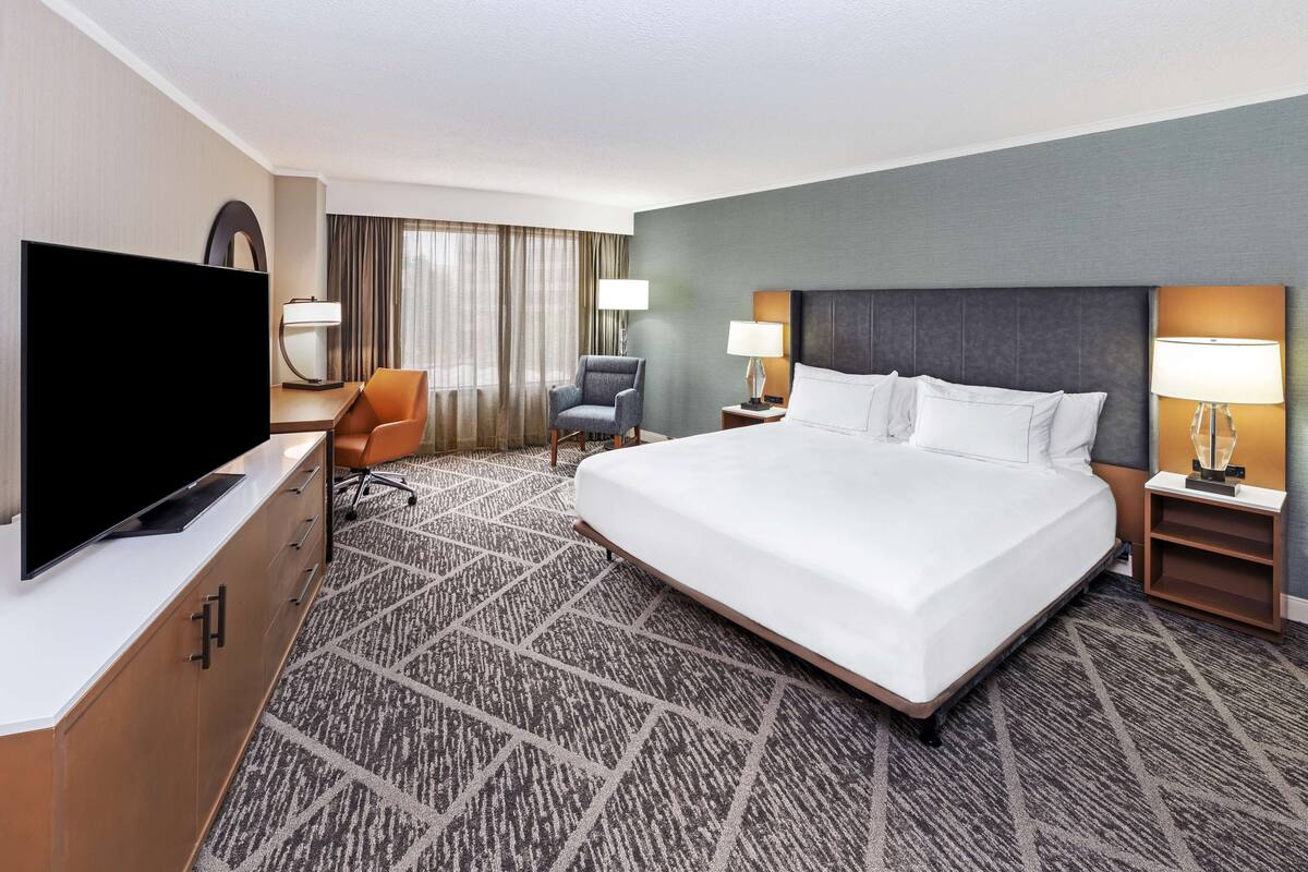 A hotel room with a large bed, a television, a desk with a chair, and a window with curtains.