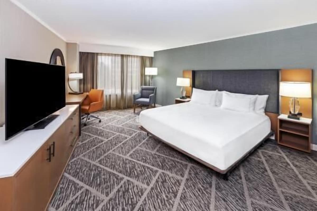 A hotel room with a large bed, a flat-screen TV, two chairs, a desk, and a nightstand with lamps.