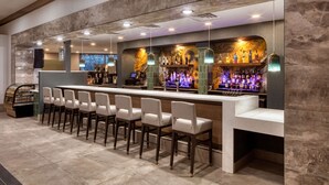 A bar area with a long counter, multiple bar stools, and a variety of liquor bottles. - DoubleTree by Hilton Tulsa - Warren Place (Tulsa)