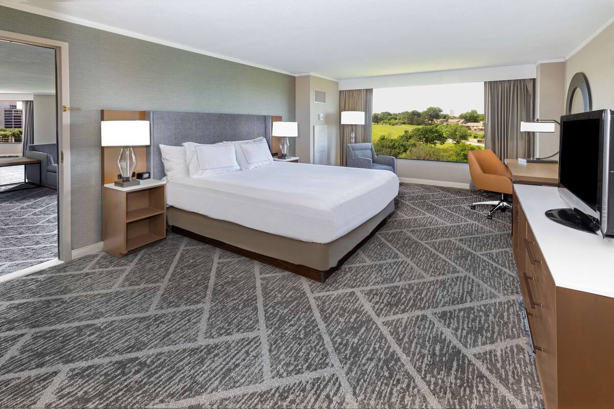 A hotel room with a large bed, two bedside tables with lamps, a desk with a flat-screen TV, and a view of the outdoors.