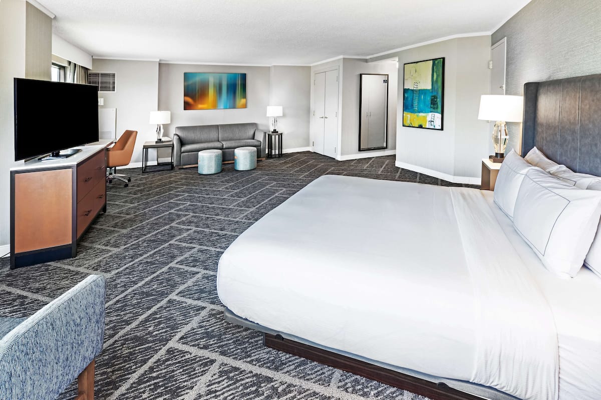 A hotel room with a large bed, a desk with a TV, a sofa, two side tables with lamps, and artwork on the walls.
