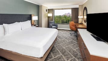 A hotel room with a large bed, a desk with a TV, a chair, and a view of trees through the window.